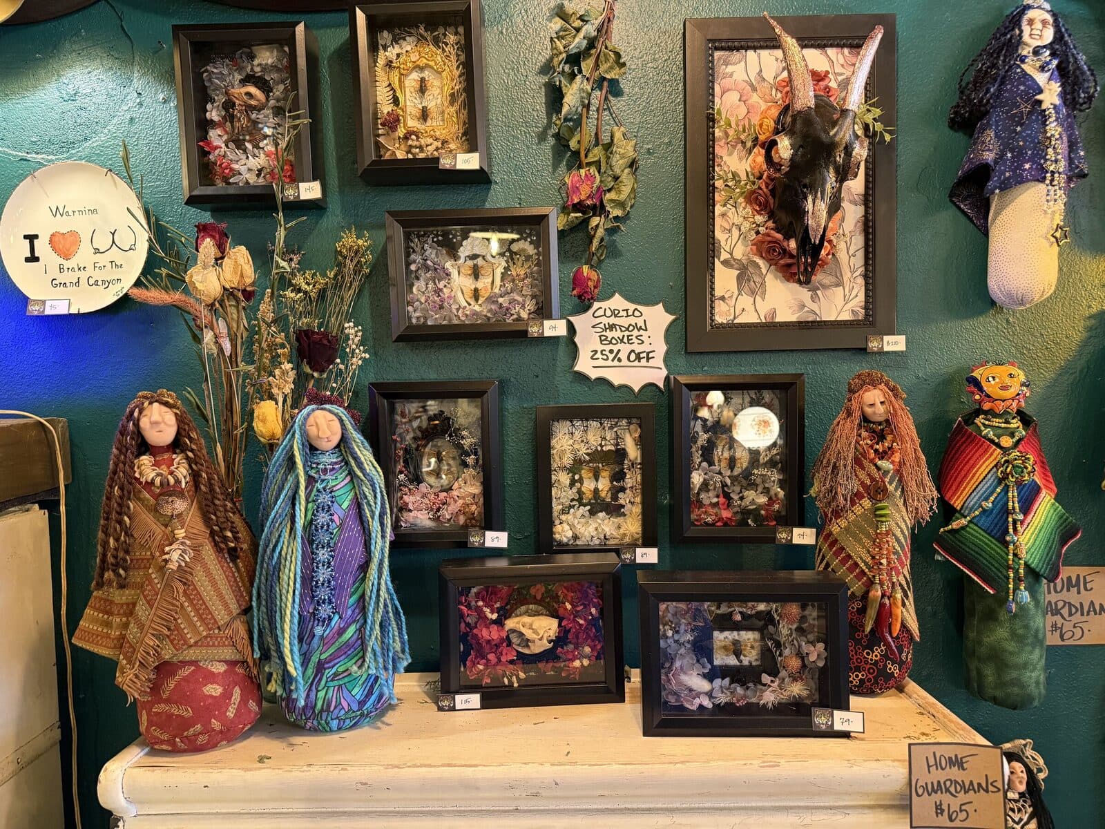 A wall of framed shadow boxes, corn-husk Saint Sage and Home Guardians dolls, and dried floral arrangements — a curated grouping in the back of the shop.