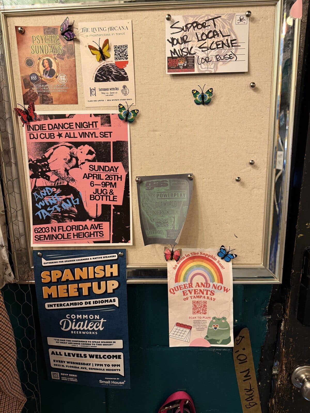 A cork bulletin board near the front door covered in flyers for local events: Indie Dance Night, Spanish Meetup, Queer and Now, Psychic Sundays, plus butterfly pin decorations.