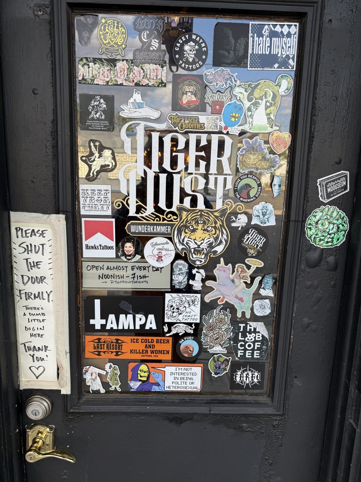 The front door of Tiger Dust: covered top to bottom in stickers from local bands, tattoo shops, and oddity shops, with a gold-and-black TIGER DUST logo dead center. A 'PLEASE SHUT THE DOOR FIRMLY' sign hangs to the left.